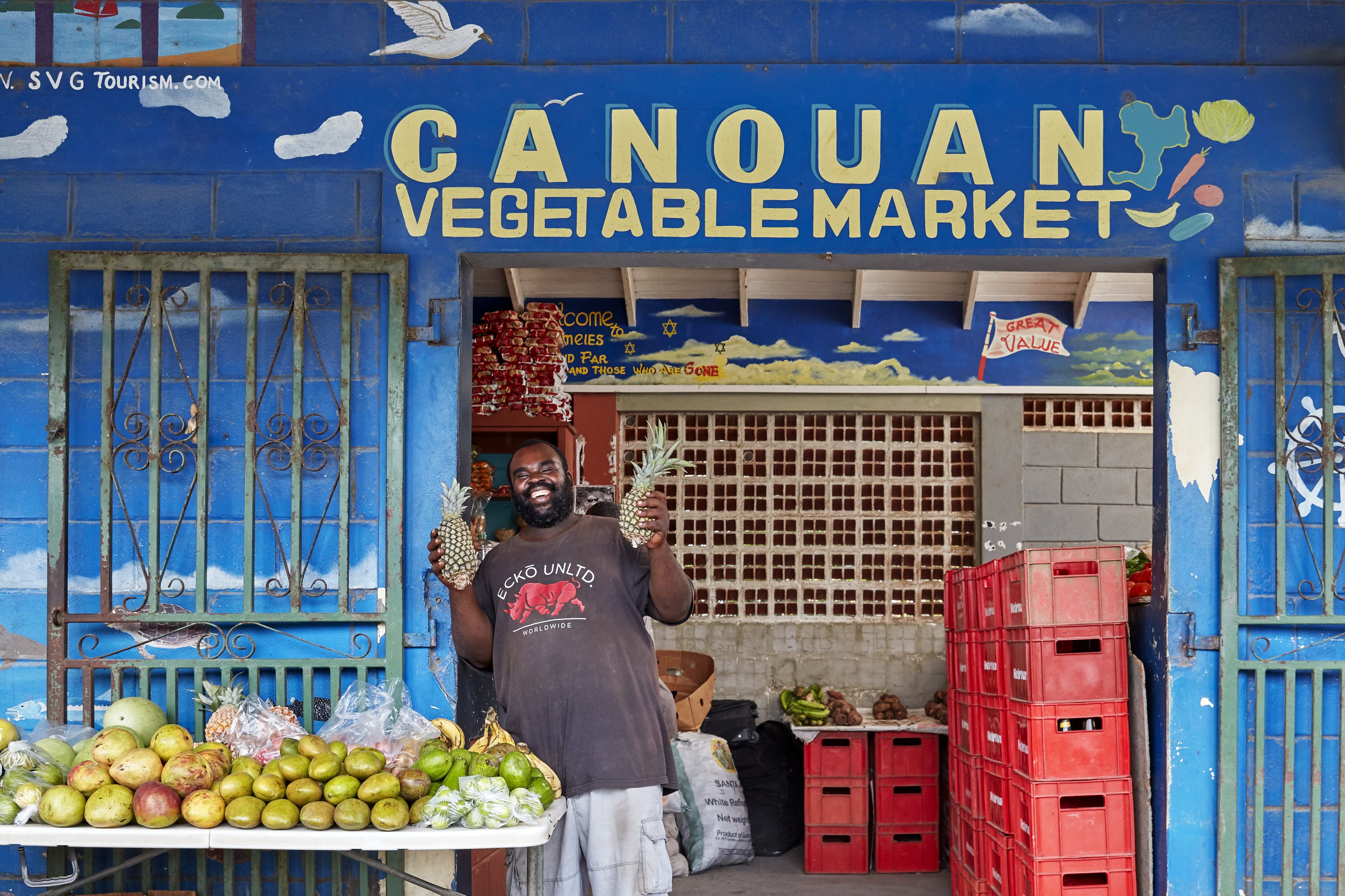 Man standing in front of a bright blue store called Canouan Vegetable Market, with a table of fruit in front of him
